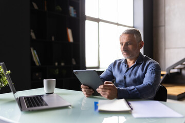 Thoughtful smiling mature businessman with tablet pc sitting in office
