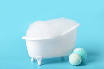 Small bathtub with foam and bath bombs on color background