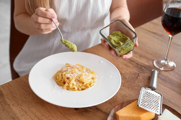 Young woman adding pesto to tasty pasta in kitchen, closeup