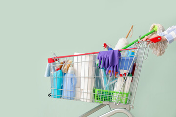 Woman with shopping cart full of cleaning supplies on pale green background