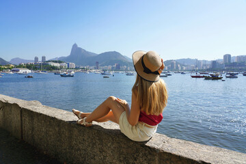 Beautiful tourist girl sitting on Mureta da Urca wall looking at stunning panoramic view of Rio de...