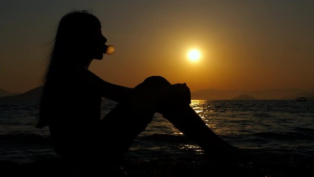 Leisure with bubble gum by sea. A teen girl silhouette have a relaxation leisure with bubble gum pn the shore against orange sun in summer.