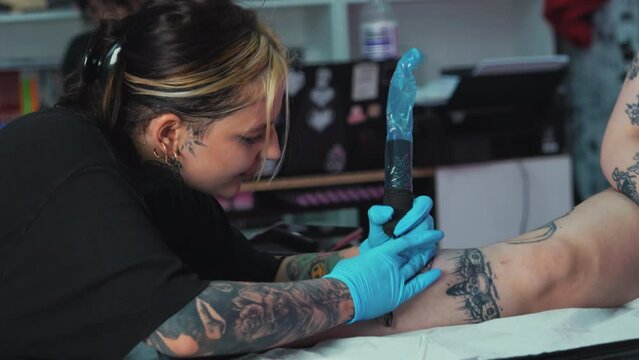 Female tattoo master doing tattoo on clients leg using a handheld tattoo machine. High quality 4k footage