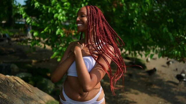 Outdoor portrait of natural Beautiful young African American woman long red braids hair style and perfect white teeth smile, posing in swimsuit at sunny summer day with green foliage beach background