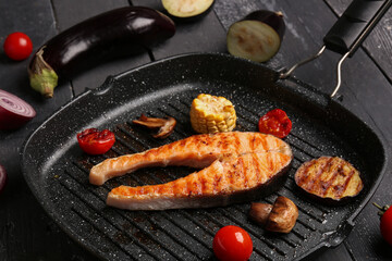 Frying pan with tasty grilled salmon steak and vegetables on black wooden background