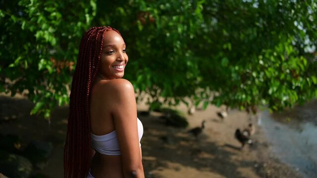 Outdoor portrait of natural Beautiful young African American woman long red braids hair style and perfect white teeth smile, posing in swimsuit at sunny summer day with green foliage beach background