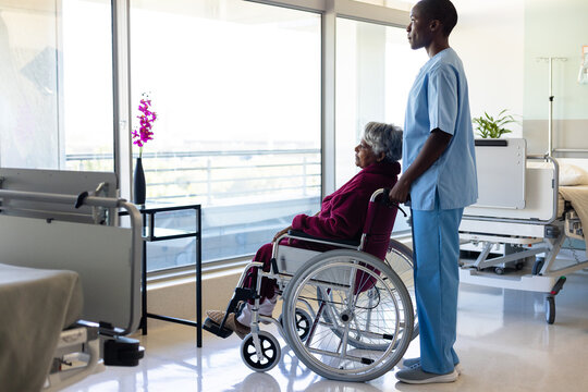Diverse female doctor and senior female patient in wheelchair, looking through window at hospital