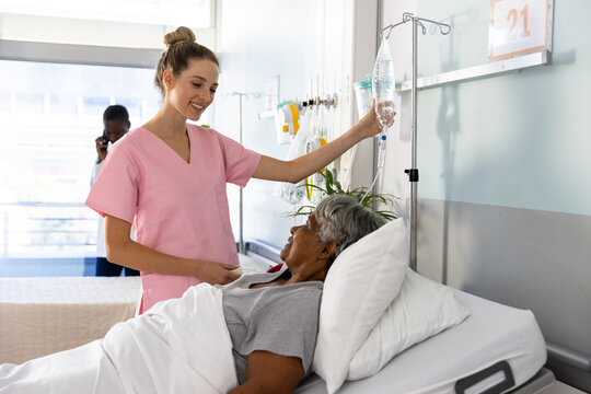 Diverse female doctor and senior female patient lying in bed, talking, holding drip at hospital