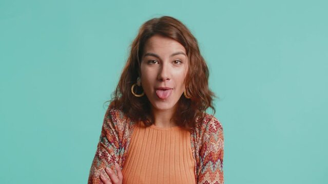 Cheerful funny young woman showing tongue making faces at camera, fooling around, joking, aping with silly face, teasing, bullying, abuse. Girl isolated alone on blue studio background. Lifestyles