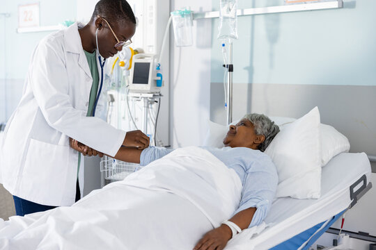 Diverse female doctor examining senior female patient lying in bed at hospital