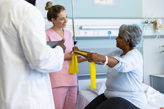 Diverse doctors and senior female patient exercising with tape at hospital