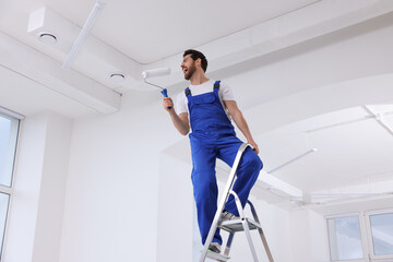 Handyman with roller having fun on step ladder in room, low angle view. Ceiling painting