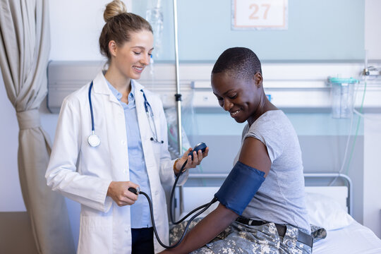Diverse female doctor examining female soldier patient, measuring blood pressure at hospital