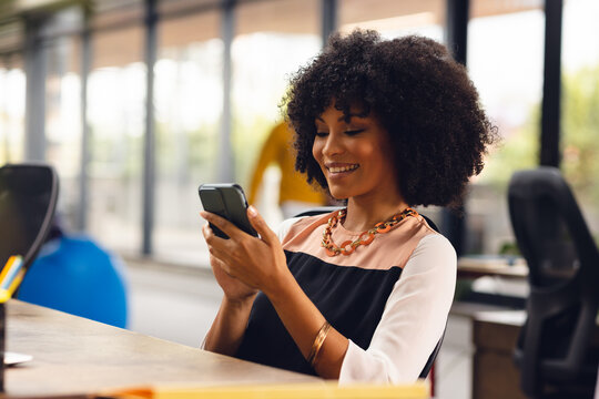 Happy african american businesswoman sitting at desk and using smartphone at office