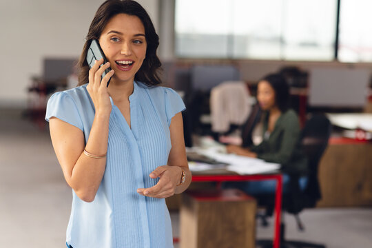 Happy Caucasian Businesswoman Talking On Smartphone, With Colleague In Background At Office