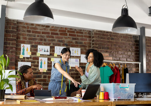 Happy diverse fashion designers using tablet and laptop, discussing project at desk in studio