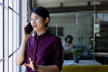 Happy asian casual businesswoman standing next to window in office talking on smartphone, copy space