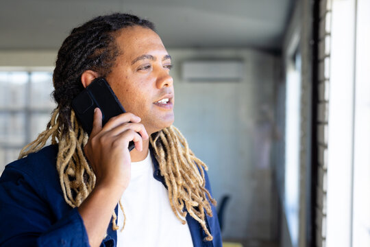 Biracial casual businessman with dreadlocks talking on smartphone standing at window in sunny office