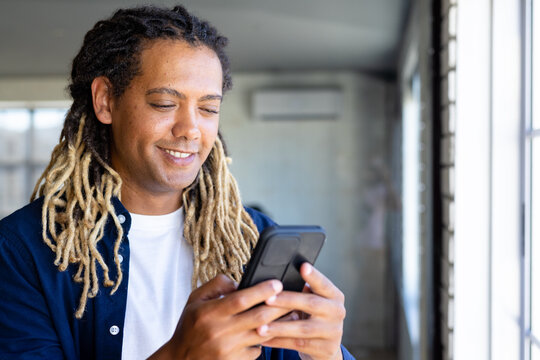 Happy biracial casual businessman with dreadlocks using smartphone by window in sunny office