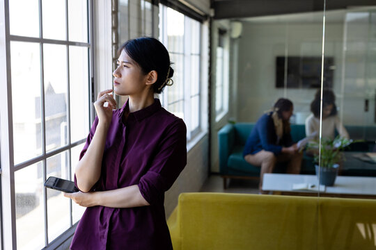 Thoughtful asian casual businesswoman standing looking out of window in office, copy space