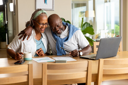 Happy senior african american couple embracing, using laptop and paying bills at home