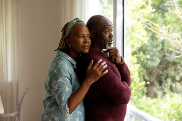 Thoughtful african american senior couple embracing, talking and looking out of window to garden