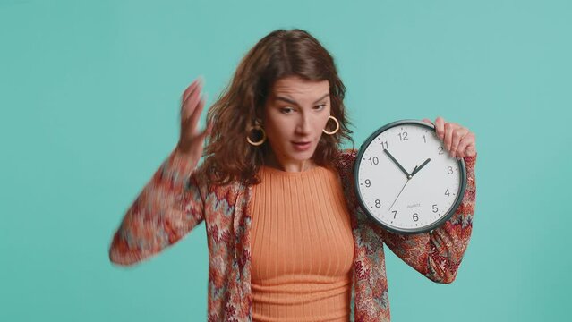 Woman with anxiety checking time on clock, running late to work, being in delay, deadline. Brunette busy girl looking at hour, minutes, worrying to be punctual isolated on blue studio background