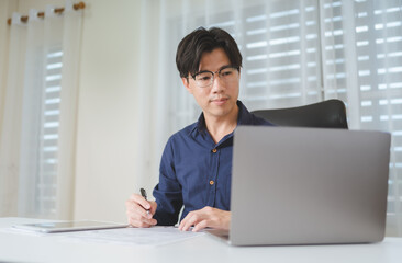 Asian Business man looking at computer laptop when planing about investment on stock market or Bitcoin Cryptocurrency, writing on paperwork for analysis and checking on financial risk and profit data. - 620362407