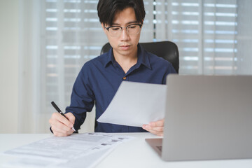 Asian Business man looking at paperwork while using computer laptop when planing about investing or trading on stock market or Bitcoin Cryptocurrency. Businessman work, analyze financial data on desk. - 620362413