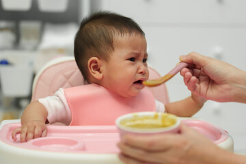 mother feeding food to her crying infant baby with spoon, refuse to eat