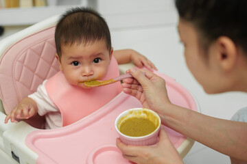 mother feeding food to her infant baby eating with spoon at home