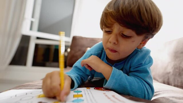 Concentrated kid drawing on paper with yellow color pen. Child laying on couch at home doing school activity. One little boy studying close up face