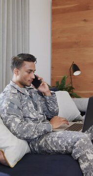 Vertical video of serious latino male soldier talking on smartphone and using laptop, slow motion