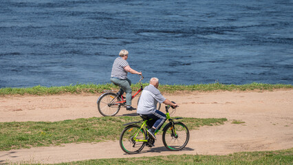 Senior couple rides bicycles along a rural road along the river bank.