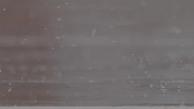 Close up shot of rain drops falling on a wooden table making splashes with chairs in the background in slowmotion LOG