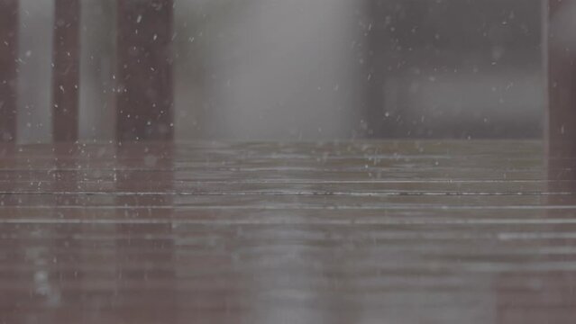 Close up shot of rain drops falling on a wooden table making splashes with chairs in the background in slowmotion LOG