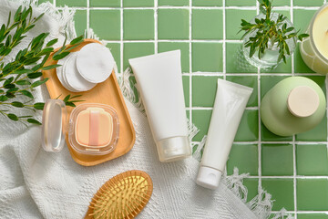 Top view of cosmetic products for hair and body care on a green tile background. White plastic tubes unbranded decorated with comb, cotton pad, cushion box and white cotton towel