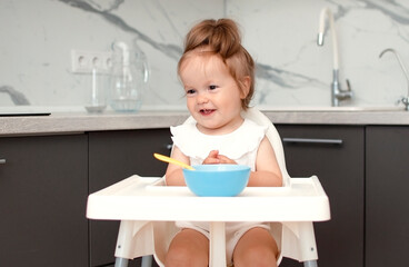 Baby is trying to eat on her own from plate, sitting on high chair. Hand with spoon for vegetable lunch. Weaning baby, first solid food for small child. Healthy baby food