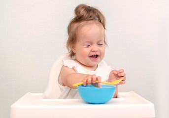 Baby is trying to eat on her own from plate, sitting on high chair. Hand with spoon for vegetable lunch. Weaning baby, first solid food for small child. Healthy baby food
