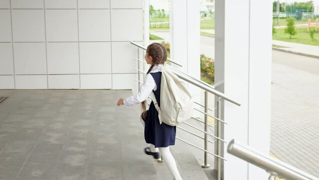 Brown-haired girl with white backpack climbs the stairs and entering primary school building. Junior schoolgirl in uniform enjoys walking alone to modern school