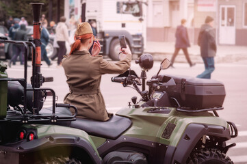 Woman at war. A young girl in military uniform on a military quad bike with a grenade launcher fixes her hair while looking at a smartphone. Morning in the city before the military parade. Back view.