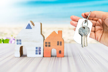 House key in girl hand with wooden house model over blurred beach background, summer beach house, buy or rent property, real estate business