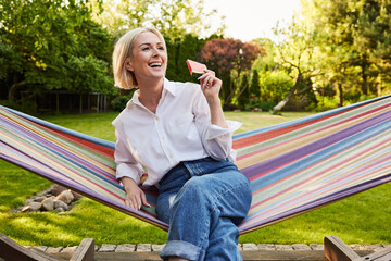 Adult woman enjoying summer afternoon in backyard garden sitting in hammock on terrace eating...