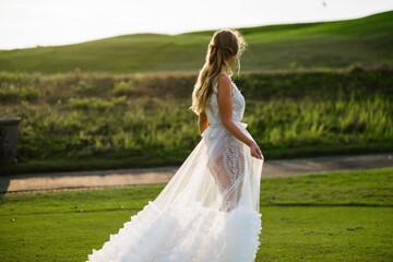 Fototapeta na wymiar A beautiful, young girl with blond hair runs across the lawn in the light of the setting sun, in a white, stylish wedding dress, rear view.