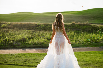 A beautiful, young girl with blond hair runs across the lawn in the light of the setting sun, in a white, stylish wedding dress, rear view.