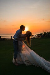  The newlyweds, in beautiful wedding clothes, hold hands and walk along the green lawn, admiring the sunset.