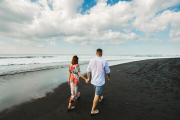 A couple of lovers, of European appearance, a man and a woman, hold hands and walk along the beach, rear view.