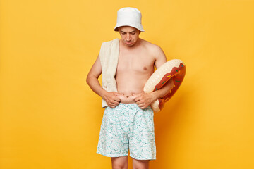 Sad unhappy adult man wearing shorts swimsuit and panama holding donut rubber ring and bag isolated...