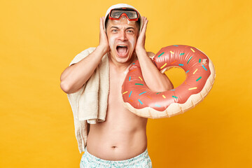 Irritated adult man wearing shorts swimsuit snorkeling goggles holding donut rubber ring and bag...