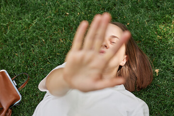 Relaxed woman wearing white t shirt lying on green grass in park outstretching hand covering her...